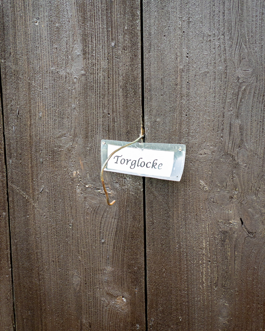 Ein hölzernes Tor an dem ein leerer Drahtbügel hängt und ein Schild mit der Aufschrift „Torglocke“ hängt.